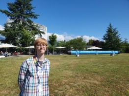 Photo of Sara Alderweireldt at CERN. She's standing on a lawn under bright blue skies with a section of beamline behind her. Lettering on the beamline reads "accelerating science" in English and French
