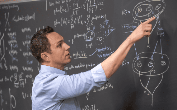 Photo of Aaron Lauda pointing at a blackboard full of writing and mathematical symbols