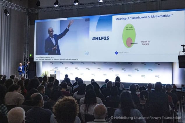 Photo of a large screen showing Sanjeev Arora gesturing at a slide. The slide is entitled 
