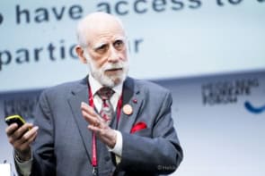 Vint Cerf speaking at the Heidelberg Laureate Forum. He's wearing a grey three-piece suit with a red tie and pocket handkerchief that matches the red conference lanyard around his neck. Behind him are words on a screen