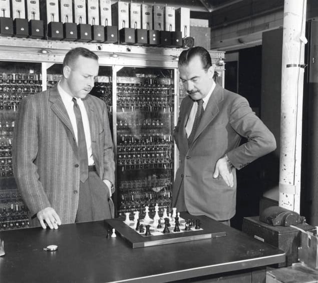 Black and white photo of two men looking at a chess board on a table in front of large rack of computer switches