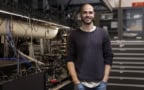 Photo of Christophe Valahu in the Quantum Control Laboratory at the University of Sydney’s Nano Institute. He's dressed casually in jeans and a black sweater with the sleeves rolled up. His hands are in his pockets and he's smiling at the camera. Behind him is an optical bench full of equipment.
