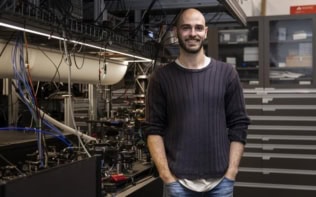 Photo of Christophe Valahu in the Quantum Control Laboratory at the University of Sydney’s Nano Institute. He's dressed casually in jeans and a black sweater with the sleeves rolled up. His hands are in his pockets and he's smiling at the camera. Behind him is an optical bench full of equipment.