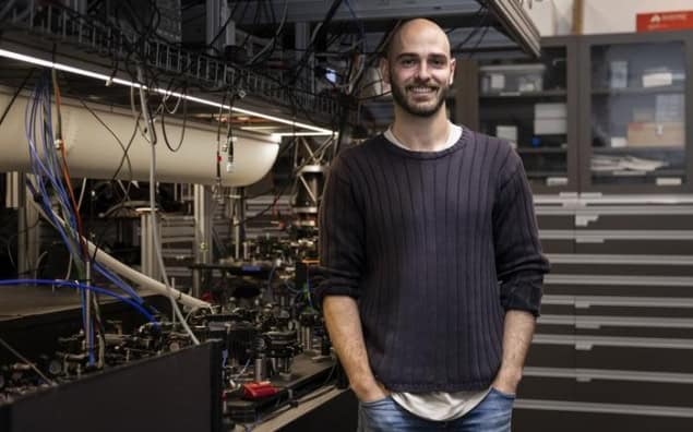 Photo of Christophe Valahu in the Quantum Control Laboratory at the University of Sydney’s Nano Institute. He's dressed casually in jeans and a black sweater with the sleeves rolled up. His hands are in his pockets and he's smiling at the camera. Behind him is an optical bench full of equipment.