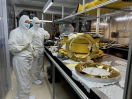 Photo of two researchers in a clean room with the components of the prototype. They're wearing white bunny suits, gloves, and masks over their noses and mouths. The components are shiny and gold and laid out on a bench
