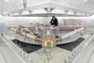 A photograph of the ring laser at the Geodetic Observatory Wettzell in Bavaria. It's mounted on a circular platform, which is itself mounted on a chunky-looking metal dais. There's a person in black clothing working on the laser at the back of the room, but most other things in the room are grey or white.