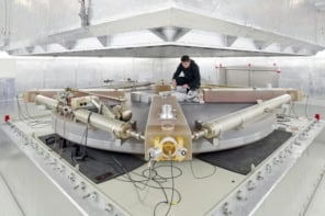 A photograph of the ring laser at the Geodetic Observatory Wettzell in Bavaria. It's mounted on a circular platform, which is itself mounted on a chunky-looking metal dais. There's a person in black clothing working on the laser at the back of the room, but most other things in the room are grey or white.