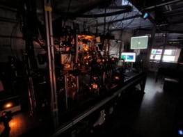 A photo of the experimental setup. It's in a laboratory with a large optical bench containing a vacuum system and optical components. Three screens in the background act as displays, and the entire scene is dark and bathed in orange laser light