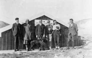 Photo of six people and a dog outside a low wooden building in a snowy landscape