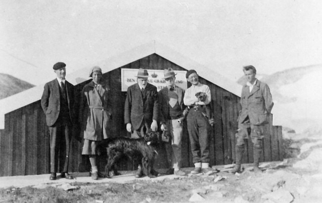 Photo of six people and a dog outside a low wooden building in a snowy landscape