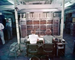 View of the backs of two women's heads as they operate a large old computer