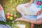 child sat cross legged on grass with a book on their lap and windfall apples by their feet