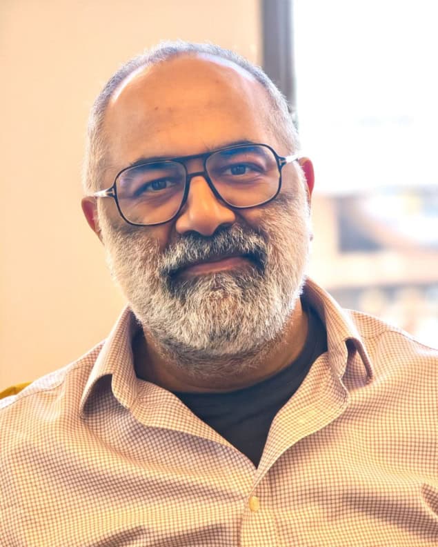 Photo of Bhavin Khatri. He has a salt-and-pepper beard and glasses, he's wearing a button-down shirt with fine red checks that's open at the collar, and he's sitting in front of a window in an office
