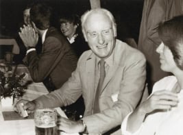 Black and white photo of people sat at a long table talking over drinks