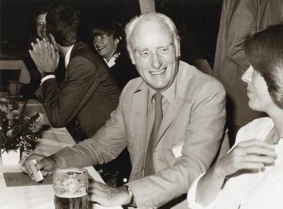 Black and white photo of people sat at a long table talking over drinks