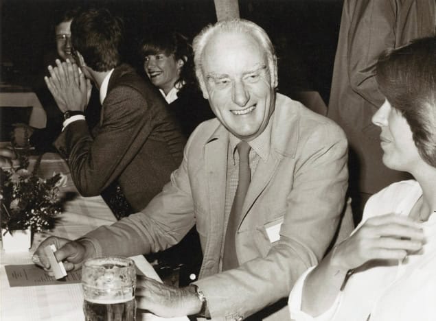 Black and white photo of people sat at a long table talking over drinks