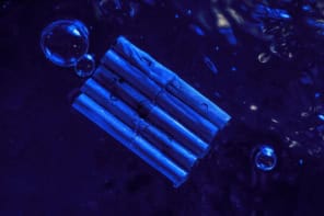 Photo showing a raft of six battered-looking metal tubes floating on the surface of a water. Everything is bathed in blue light and a few bubbles are present.