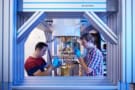 Photo of two people standing either side of dilution refrigerator, taken through an aperture in the refrigerator's supporting frame. They're wearing blue nitrile gloves and working on the refrigerator's connections.