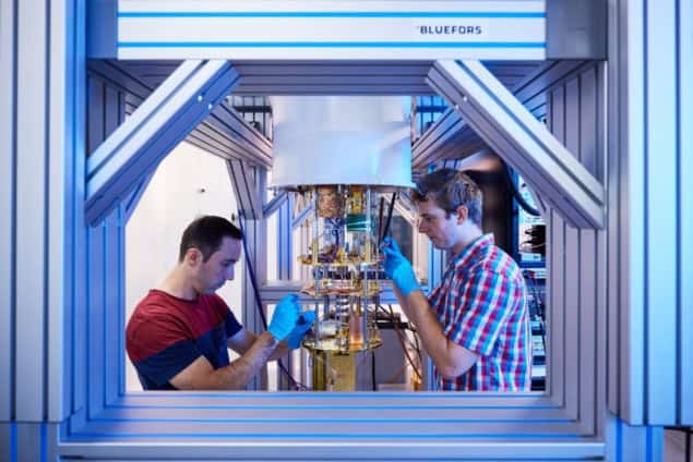 Photo of two people standing either side of dilution refrigerator, taken through an aperture in the refrigerator's supporting frame. They're wearing blue nitrile gloves and working on the refrigerator's connections.