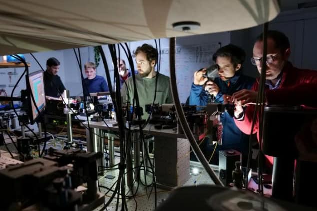 A group of physicists gathered around an optical bench in a laboratory. They are variously studying screens, looking through an IR viewer, and adjusting equipment on the bench.