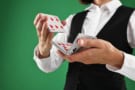 Close-up image of a woman's hands shuffling a deck of cards. She's wearing a black waistcoat and a crisp white shirtand standing against a green background. Her face isn't in the frame.