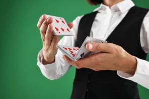 Close-up image of a woman's hands shuffling a deck of cards. She's wearing a black waistcoat and a crisp white shirtand standing against a green background. Her face isn't in the frame.
