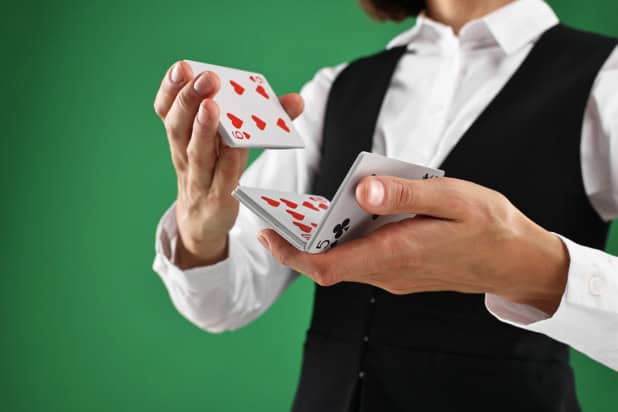 Close-up image of a woman's hands shuffling a deck of cards. She's wearing a black waistcoat and a crisp white shirtand standing against a green background. Her face isn't in the frame.