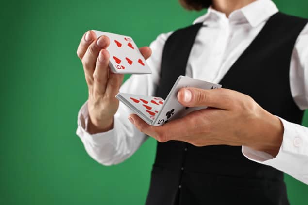 Close-up image of a woman's hands shuffling a deck of cards. She's wearing a black waistcoat and a crisp white shirtand standing against a green background. Her face isn't in the frame.
