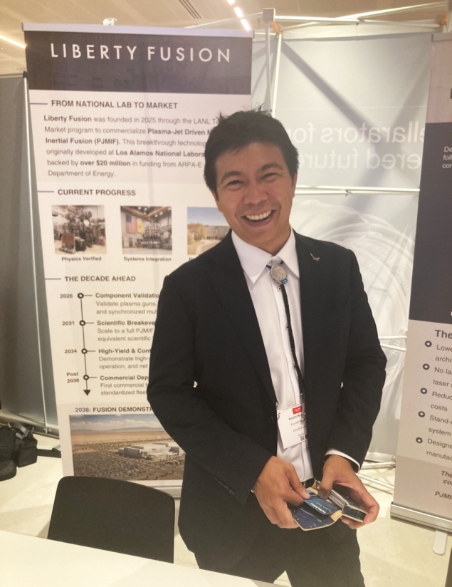 Photo of Koichi Masuda standing in front of a poster for his company, Liberty Fusion, at Fusion Fest in London. He's wearing a suit with a bolo tie, the sartorial symbol of Los Alamos.
