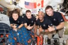 Four astronauts floating in a space capsule holding thumbs up and smiling