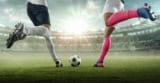 Two football players converging on the ball with legs raised ready to kick