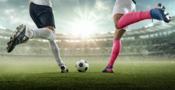 Two football players converging on the ball with legs raised ready to kick