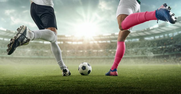Two football players converging on the ball with legs raised ready to kick
