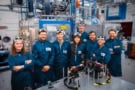 Photo of several researchers in a laboratory. They're all wearing identical dark blue shirts and have laser safety goggles on their heads