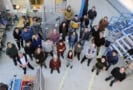 Photo of 28 researchers in the collaboration standing on the experimental floor at GSI/FAIR. The researchers are all dressed casually and a set of metal stairs and other equipment are visible in the background. The photo is taken from above and everyone is looking up at the camera.