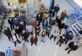 Photo of 28 researchers in the collaboration standing on the experimental floor at GSI/FAIR. The researchers are all dressed casually and a set of metal stairs and other equipment are visible in the background. The photo is taken from above and everyone is looking up at the camera.