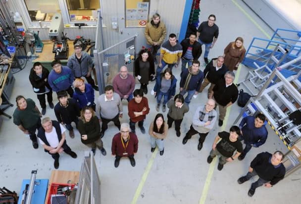 Photo of 28 researchers in the collaboration standing on the experimental floor at GSI/FAIR. The researchers are all dressed casually and a set of metal stairs and other equipment are visible in the background. The photo is taken from above and everyone is looking up at the camera.