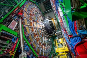 Photo of the CMS detector, which resembles a multicoloured wheel held up by green steel beams. A yellow lift vehicle is visible underneath it.