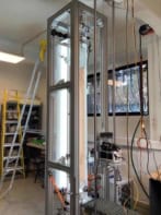 Photo of the experimental apparatus in the laboratory, showing a tall, well-lit transparent box supported by a metal frame. A camera is mounted on a vertical track next to the box and there are ladders in the background.