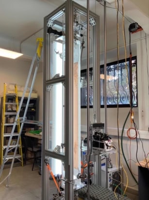 Photo of the experimental apparatus in the laboratory, showing a tall, well-lit transparent box supported by a metal frame. A camera is mounted on a vertical track next to the box and there are ladders in the background.