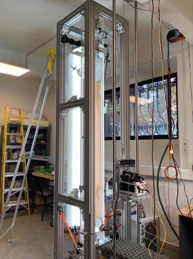 Photo of the experimental apparatus in the laboratory, showing a tall, well-lit transparent box supported by a metal frame. A camera is mounted on a vertical track next to the box and there are ladders in the background.