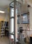 Photo of the experimental apparatus in the laboratory, showing a tall, well-lit transparent box supported by a metal frame. A camera is mounted on a vertical track next to the box and there are ladders in the background.
