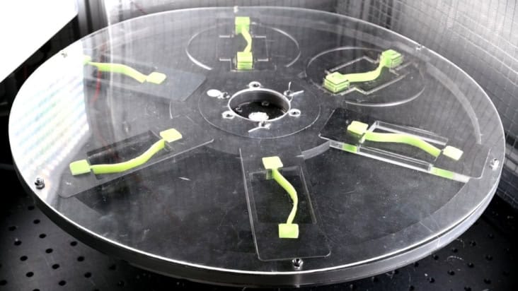 A still from a video showing six flexible strips of material on a wheel. When the wheel spins, the strips of material snap into left-bent or right-bent configurations depending on the dynamics of the spin.