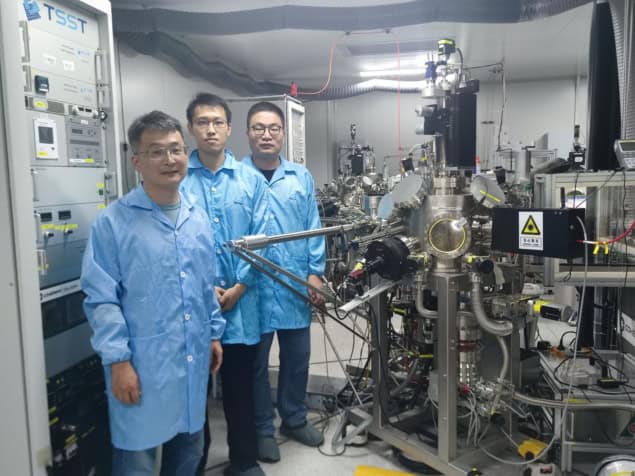 Three men standing in a laboratory next to a rack of equipment and a vacuum chamber with many ports and hoses. The men are wearing blue lab coats and protective booties over their shoes.