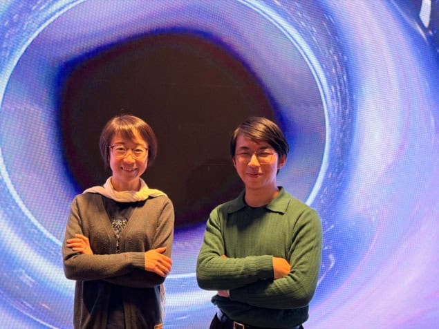Photo of Ling Sun and Neil Lu standing in front of a glowing pixelated backdrop depicting a black hole surrounded by a swirl of blue and purple