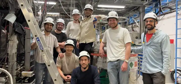 Photo of eight members of the ALPHA collaboration in the lab at CERN. They're all wearing white hard hats and are casually dressed in jeans and t-shirts. A ladder leans against an experimental apparatus in the foreground and a tank of liquid helium is visible in the background, as are lots of cables.