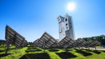 Photo of a solar tower power plant. A green field full of mirrors directs sunlight onto a tower under a blue sky and a white Sun