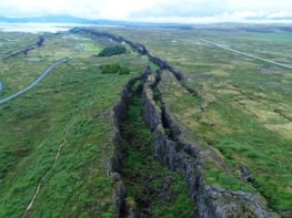 Aerial photo of a green landscape with a ragged gash of black, exposed rock running through it. A couple of roads and paths run parallel to the gash and a lake and mountains are visible in the background.
