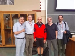 A group of five physicists standing in a lecture theatre in front of a display cabinet and a large blackboard.