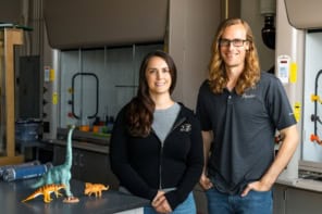 Photo of Alexandria Johnson and Brandon Johnson in a laboratory. They're casually dressed and there are some model dinosaurs sitting on a lab bench next to them. There's a fume hood in the background.